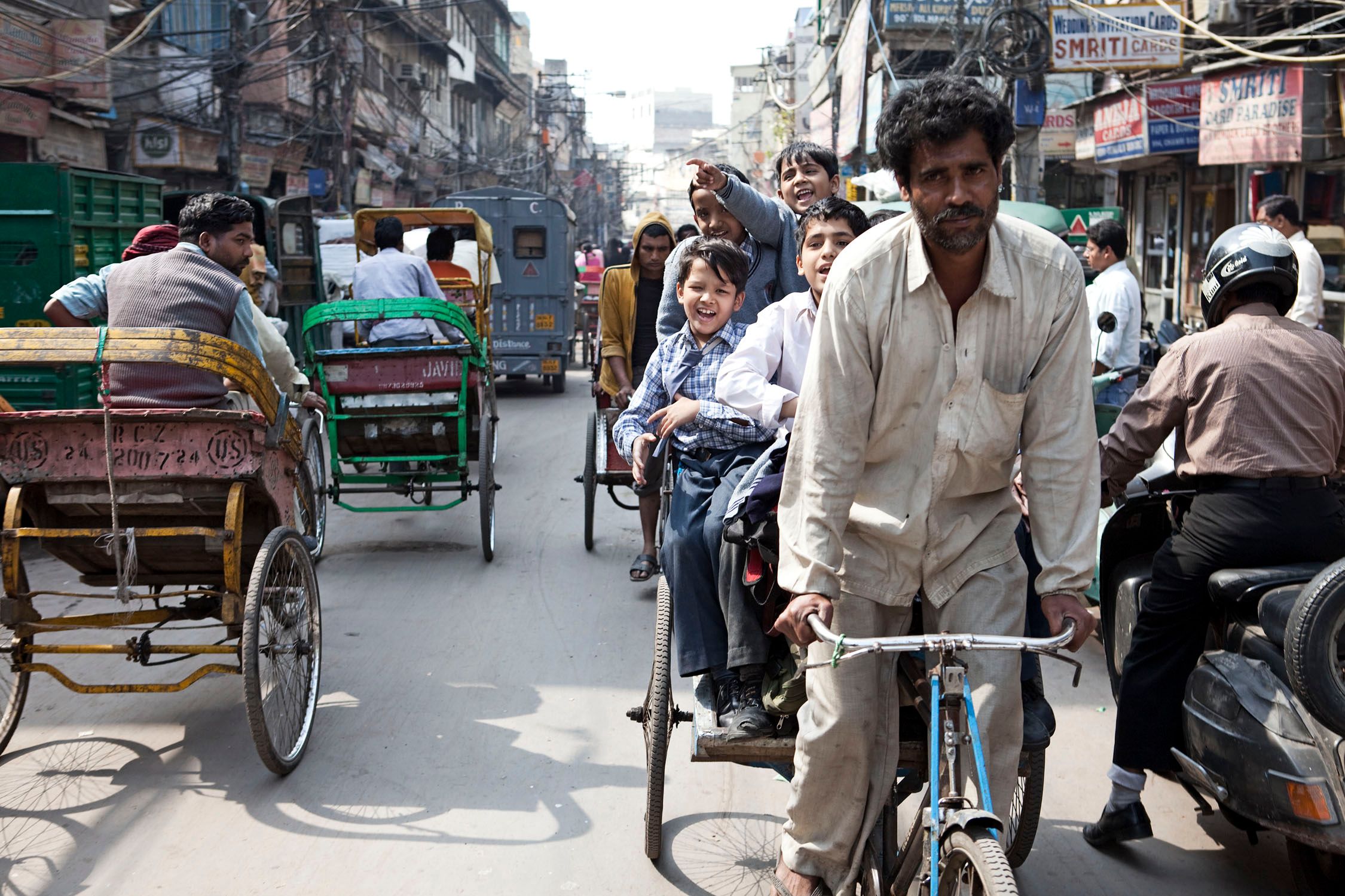 A crowded street in India with a man on a rickshaw, a boy on a rickshaw, and a man on a motorcycle, all surrounded by other rickshaws and people.