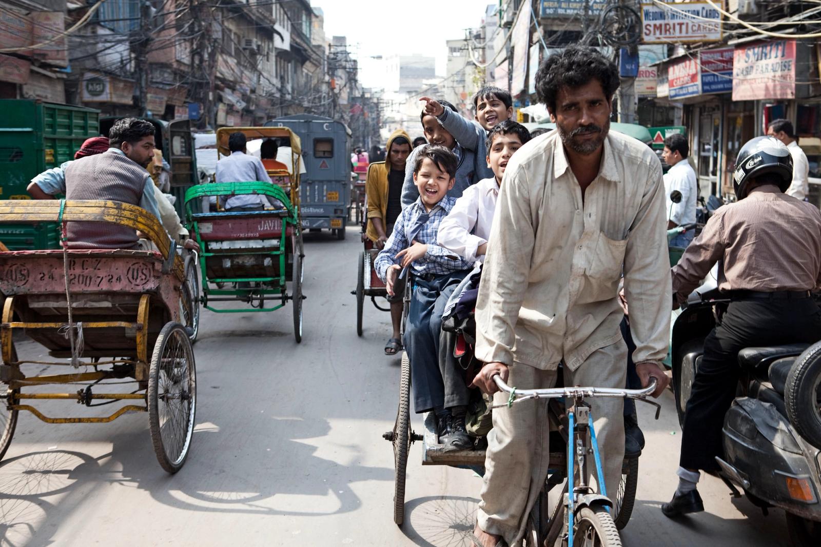 A crowded street in India with a man on a rickshaw, a boy on a rickshaw, and a man on a motorcycle, all surrounded by other rickshaws and people.