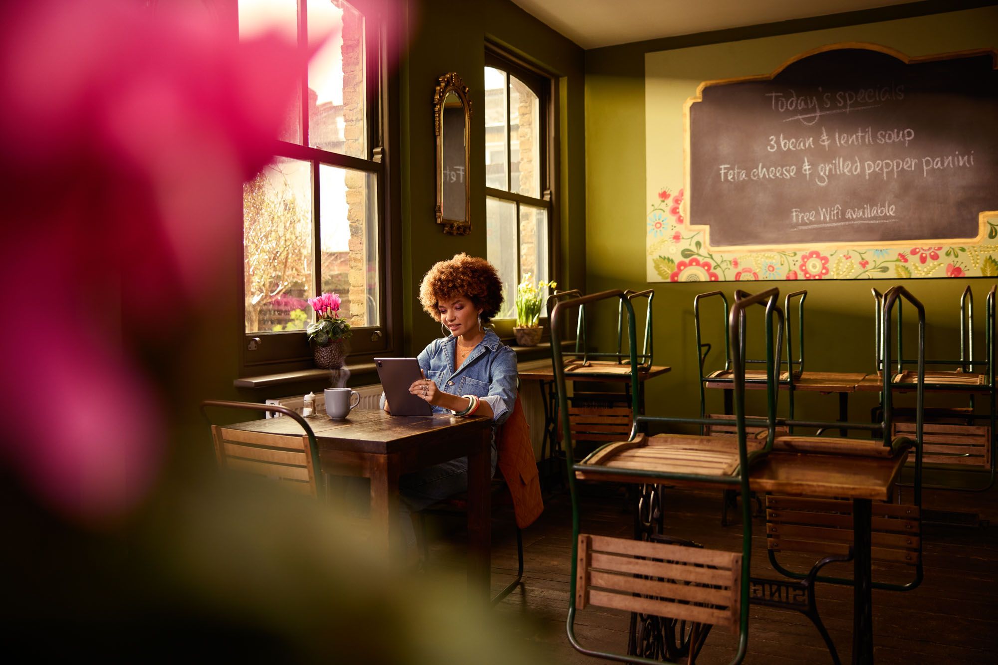 A woman with curly hair and a denim jacket sits at a table in a cafe, working on a laptop.
