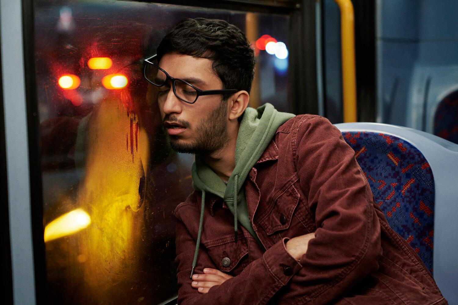 A man with dark hair and a beard, wearing a red jacket and a green hoodie, sitting on a bus at night, looking out the window at the city lights.