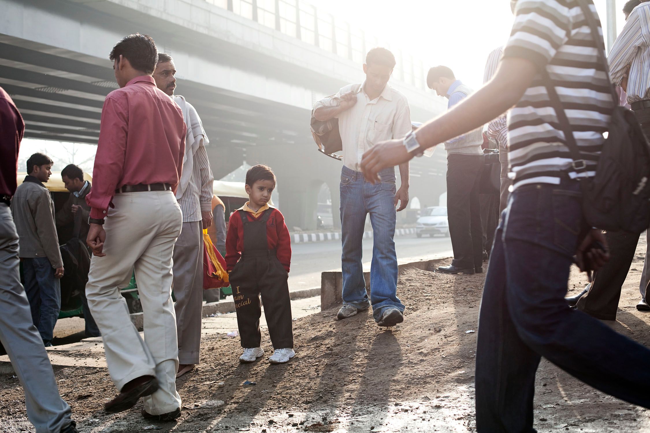 A young boy stands in a crowded street with adults and children, near a large bridge.