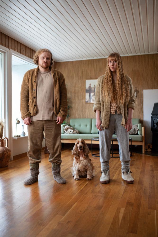 A man and a woman, both with long hair, standing in a living room with a dog sitting between them.