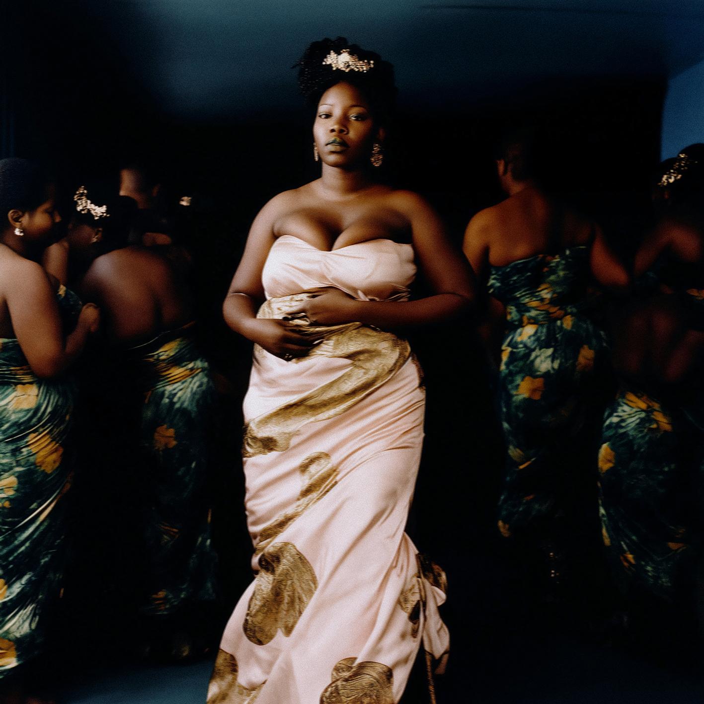 A woman in a white, strapless evening gown stands in front of a dark background, surrounded by identical dark-skinned women in the same gowns.