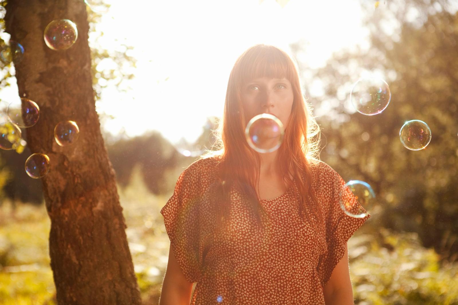 A woman in a red and white polka dot dress stands in a forest with bubbles around her.