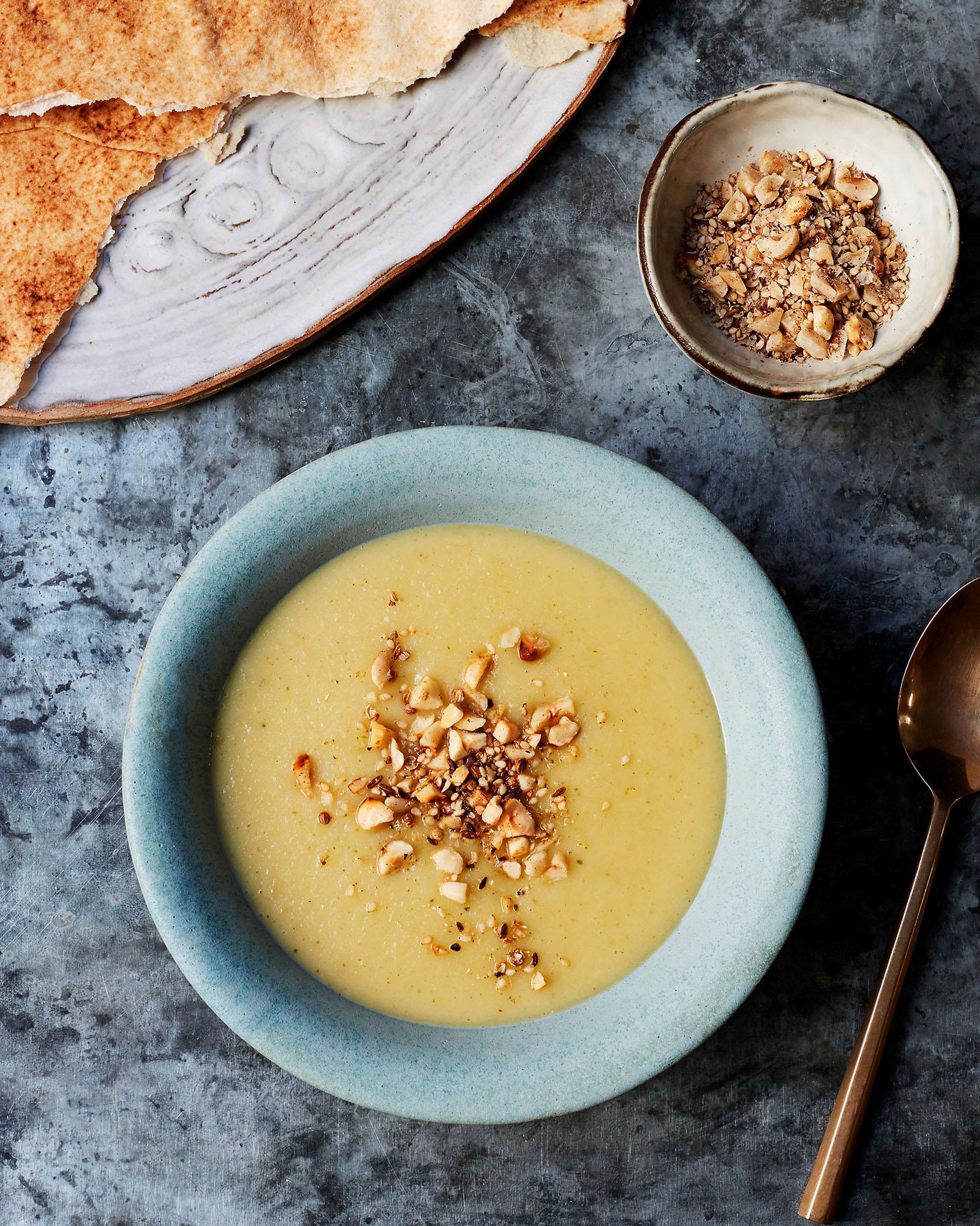 A bowl of creamy yellow soup with chopped nuts and pita bread on the side.