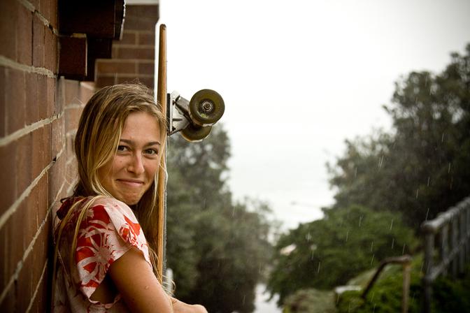 A young woman with long blonde hair and a red and white striped shirt is standing in the rain, holding a skateboard.