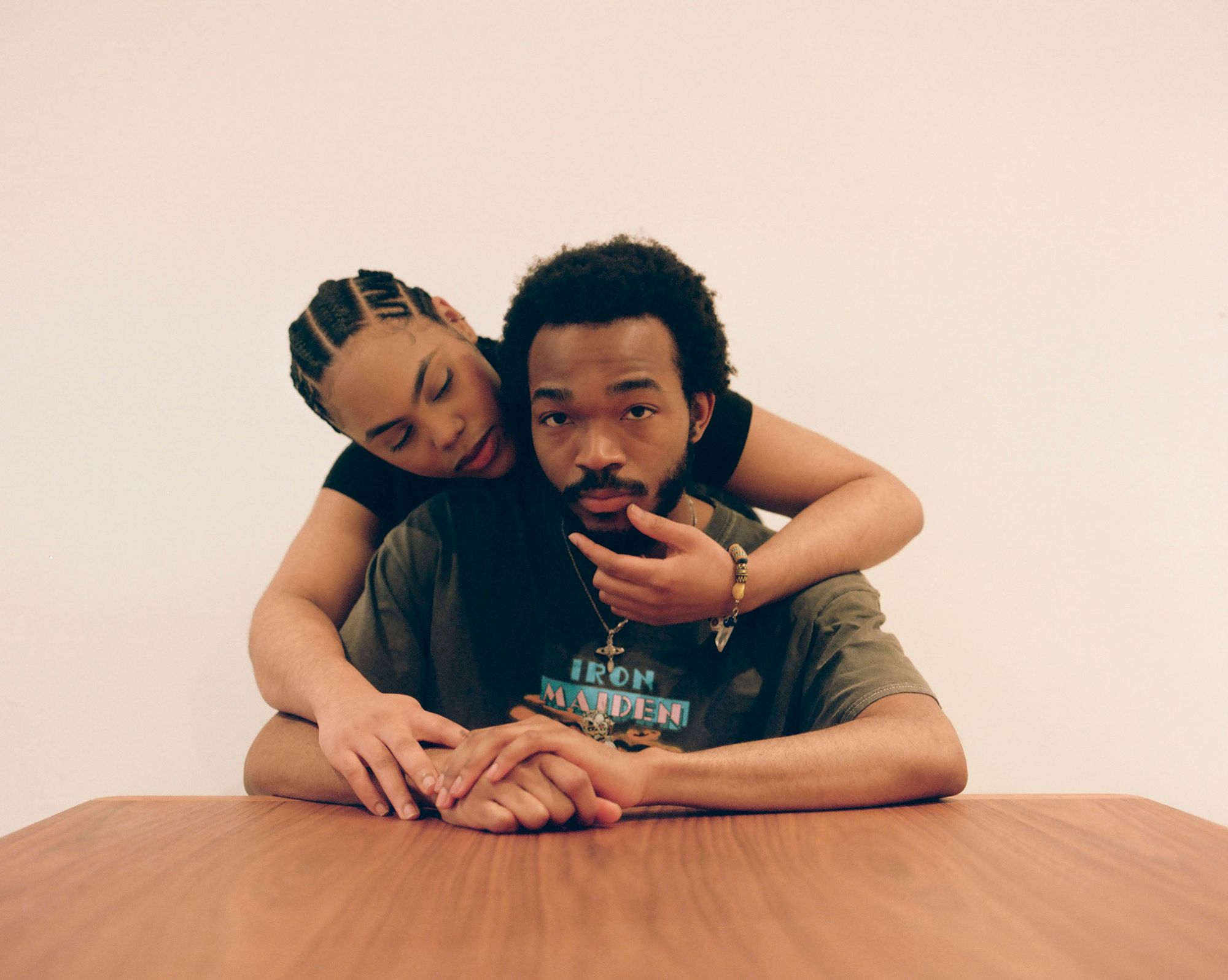 A man and a woman sitting at a table, with the woman leaning over the man's shoulder and her hand on his face.