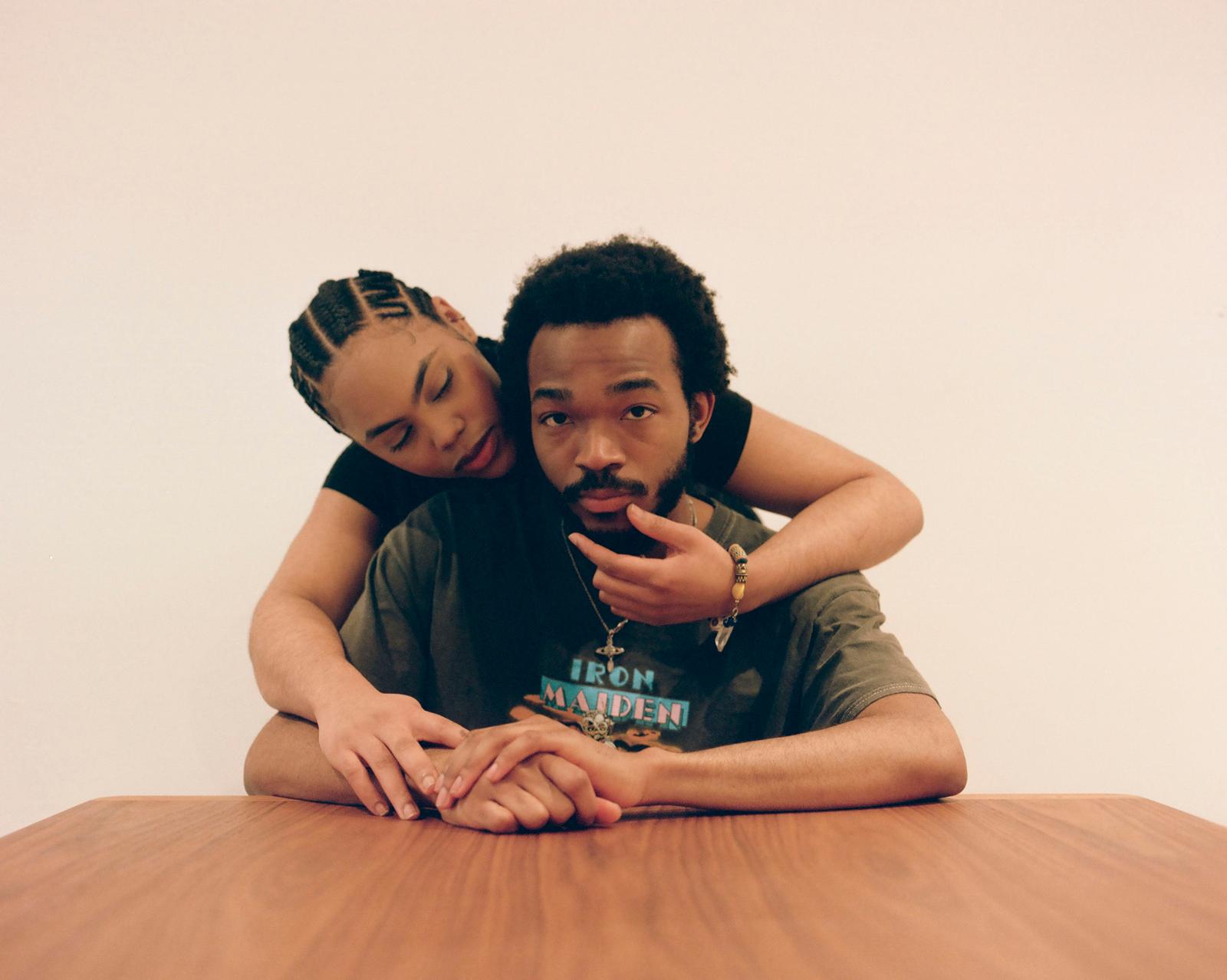 A man and a woman sitting at a table, with the woman leaning over the man's shoulder and her hand on his face.