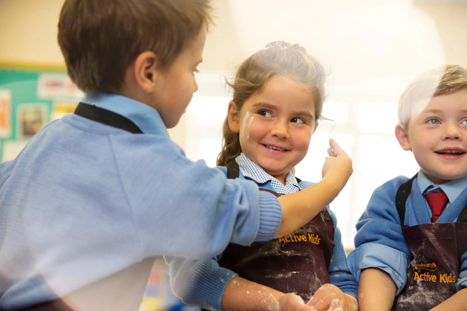 Three children in school uniforms are playing with a white powder, possibly flour or face paint, in a classroom setting.
