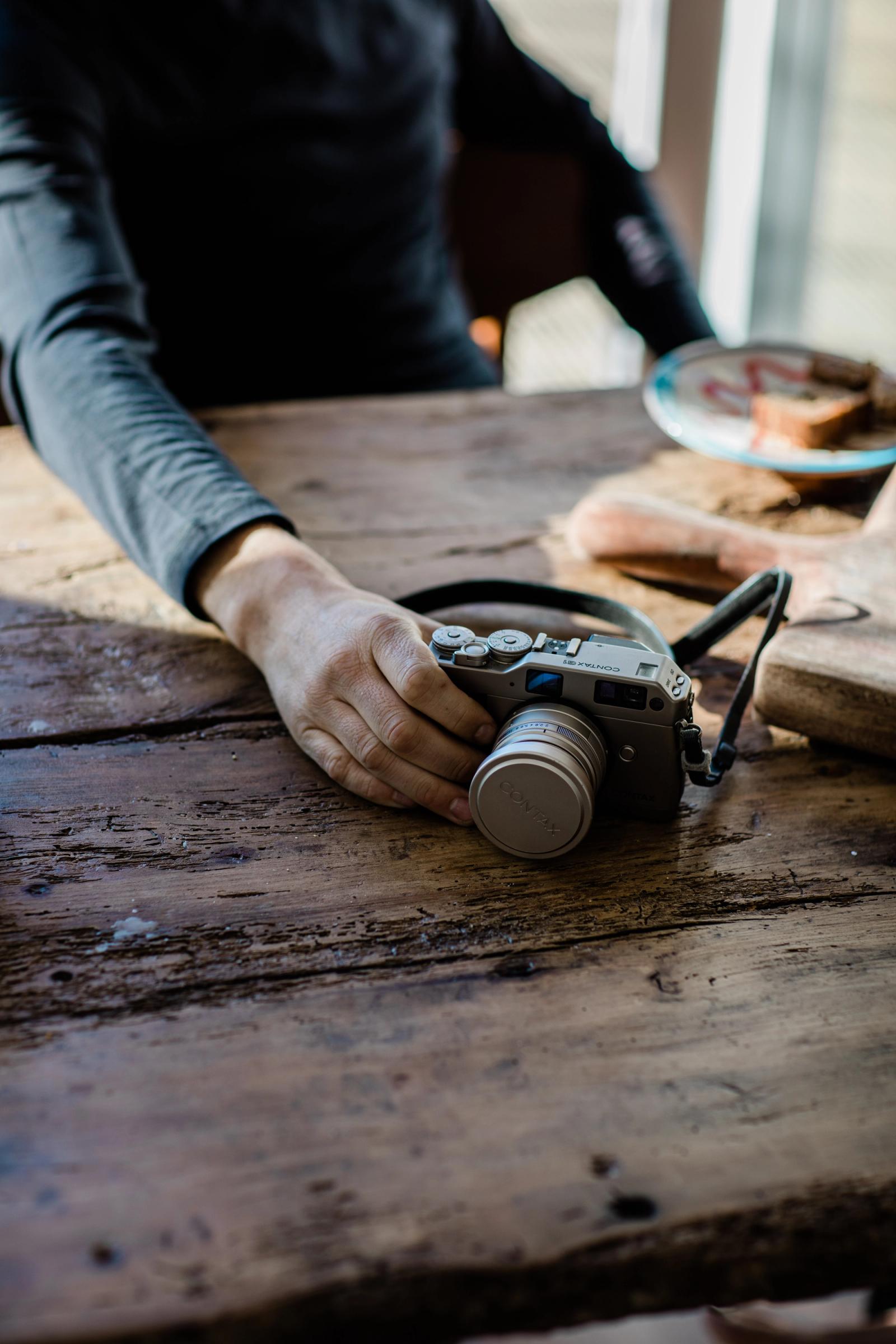 A person's hand is holding a camera on a wooden table, with a plate of food in the background.