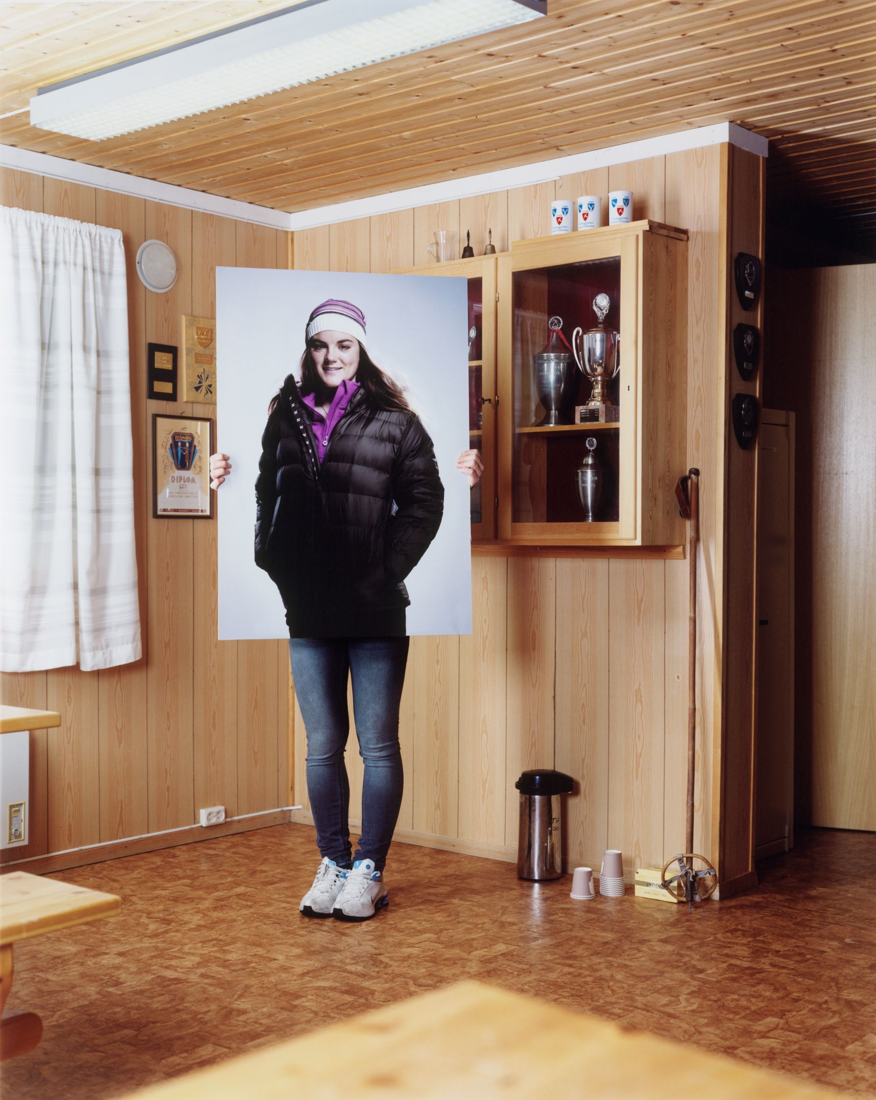 A woman in a black puffer jacket, jeans, and sneakers, standing in a room with a wooden wall and a few sports-related items, such as a hockey stick and a soccer ball.