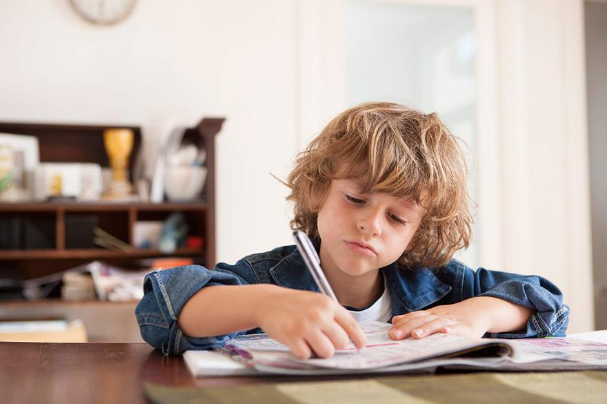 A young boy with messy, curly brown hair is sitting at a table, wearing a denim jacket and a white t-shirt, intently coloring in a coloring book with a silver pen.