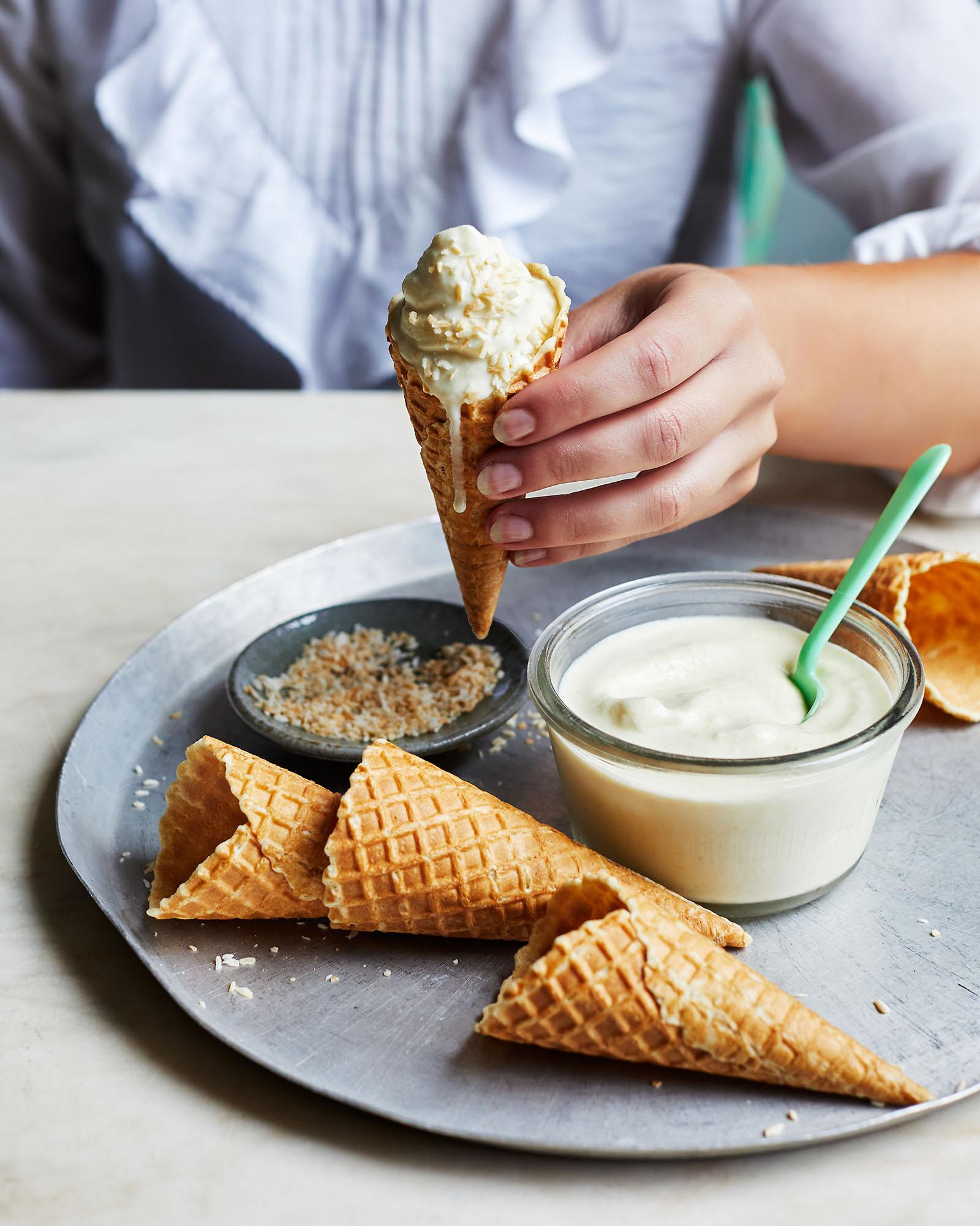 A person is holding a cone of ice cream, with a few cones and a bowl of ice cream in the background.