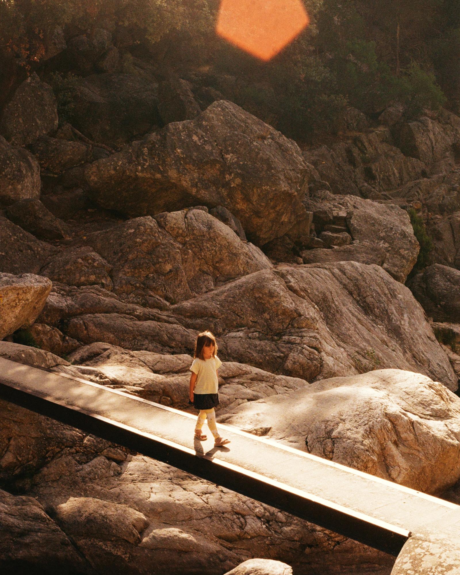 A young girl in a white t-shirt and black skirt walking on a narrow, elevated path surrounded by large rocks and boulders, with a forest or mountainous landscape in the background.