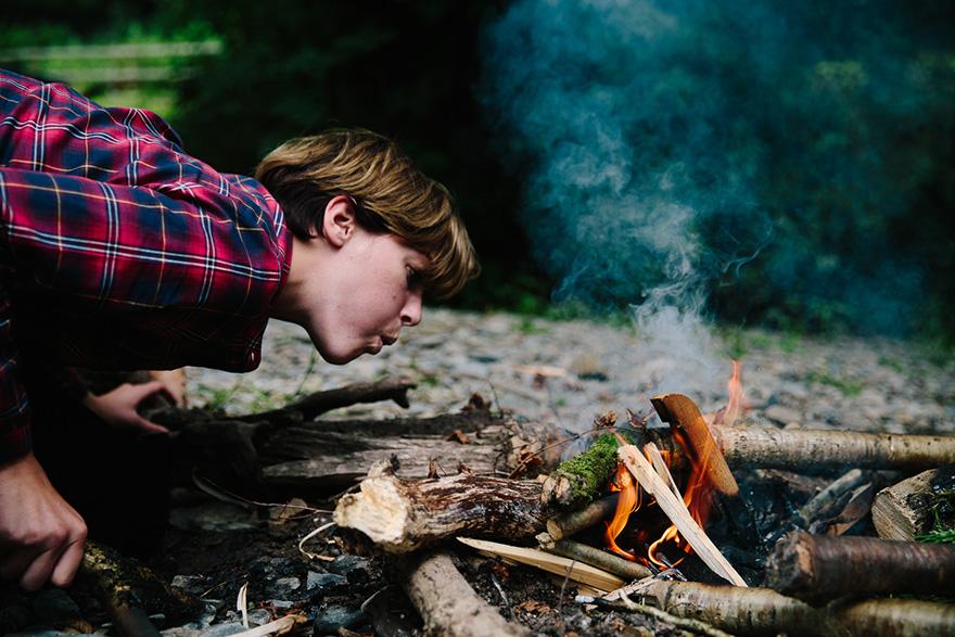 A person in a red plaid shirt and jeans is crouched over a small fire in a forest, surrounded by fallen trees and branches.