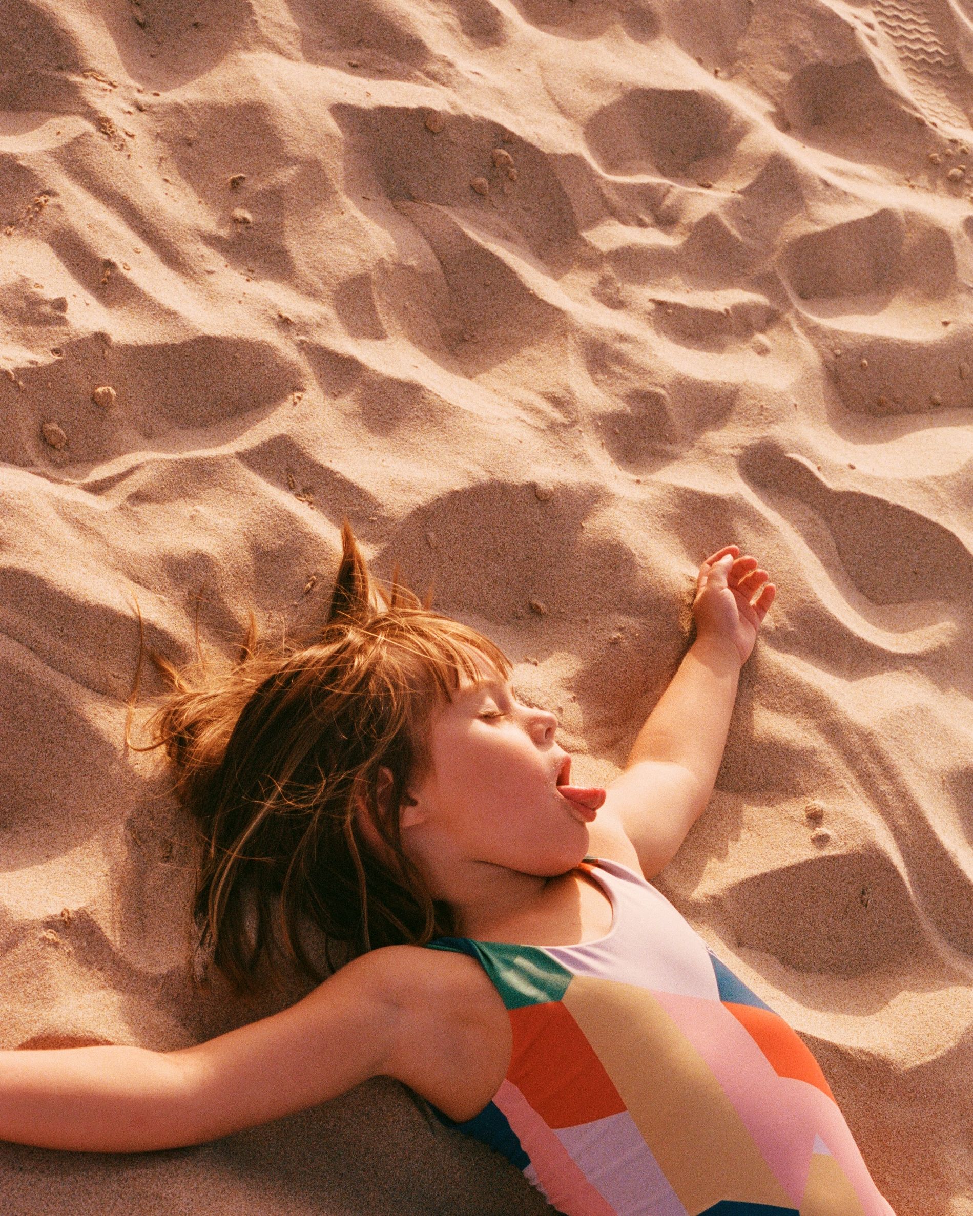 A young girl in a colorful swimsuit is lying on her back in the sand, with her arms stretched out to the sides and her eyes closed, enjoying the warm sun on her skin.