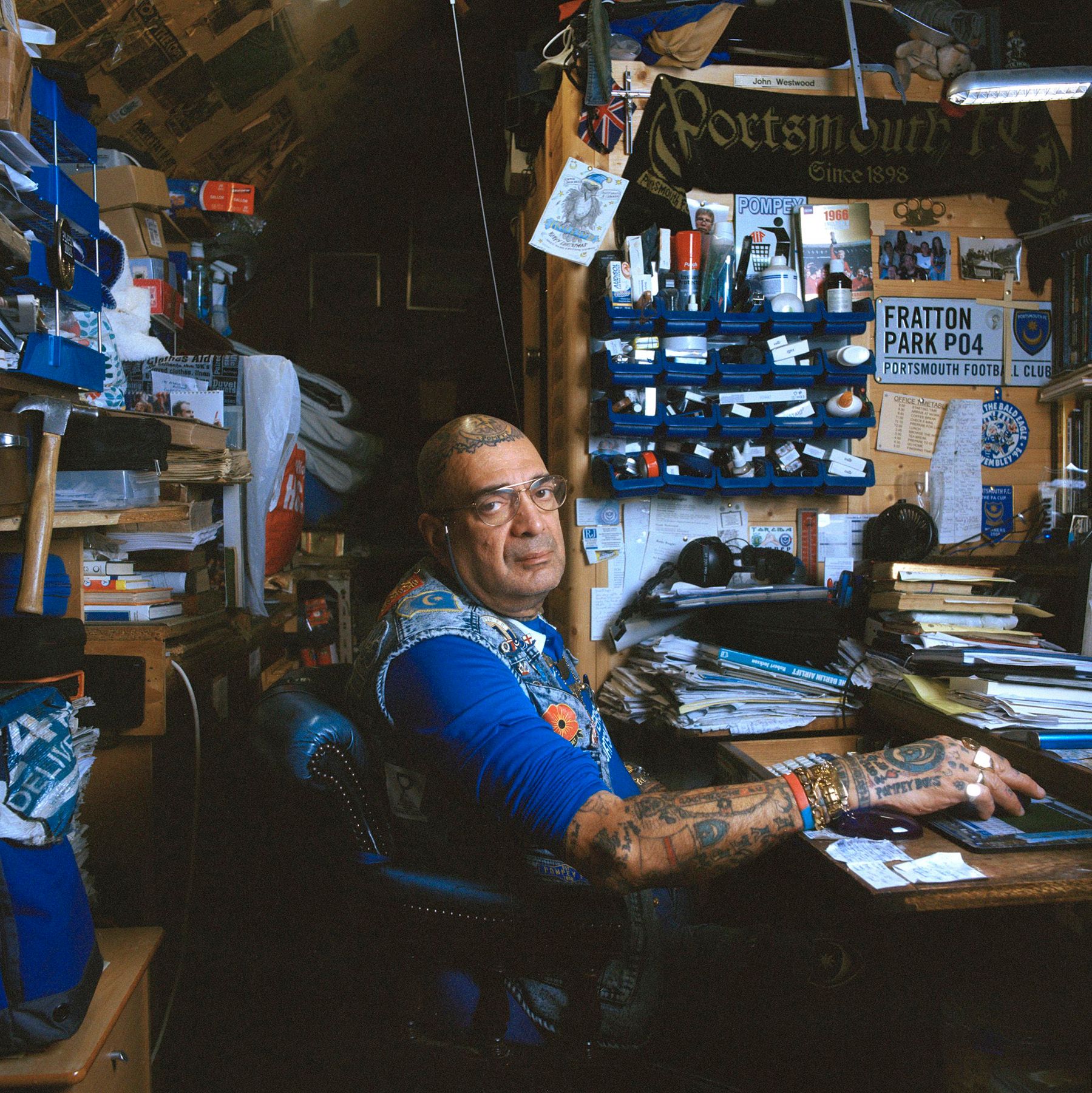 A man sits at a cluttered desk in a dimly lit room, surrounded by papers, books, and objects.