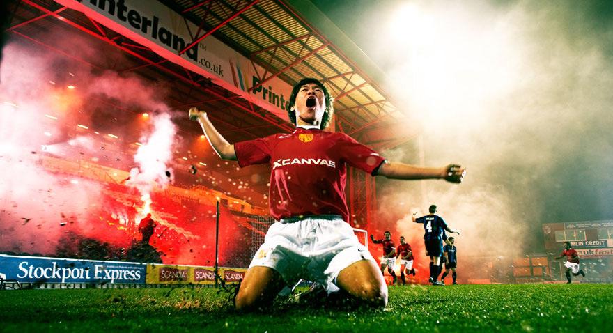 A football player in a red jersey and white shorts celebrates a goal in a stadium surrounded by fans, with a large screen displaying a football match.