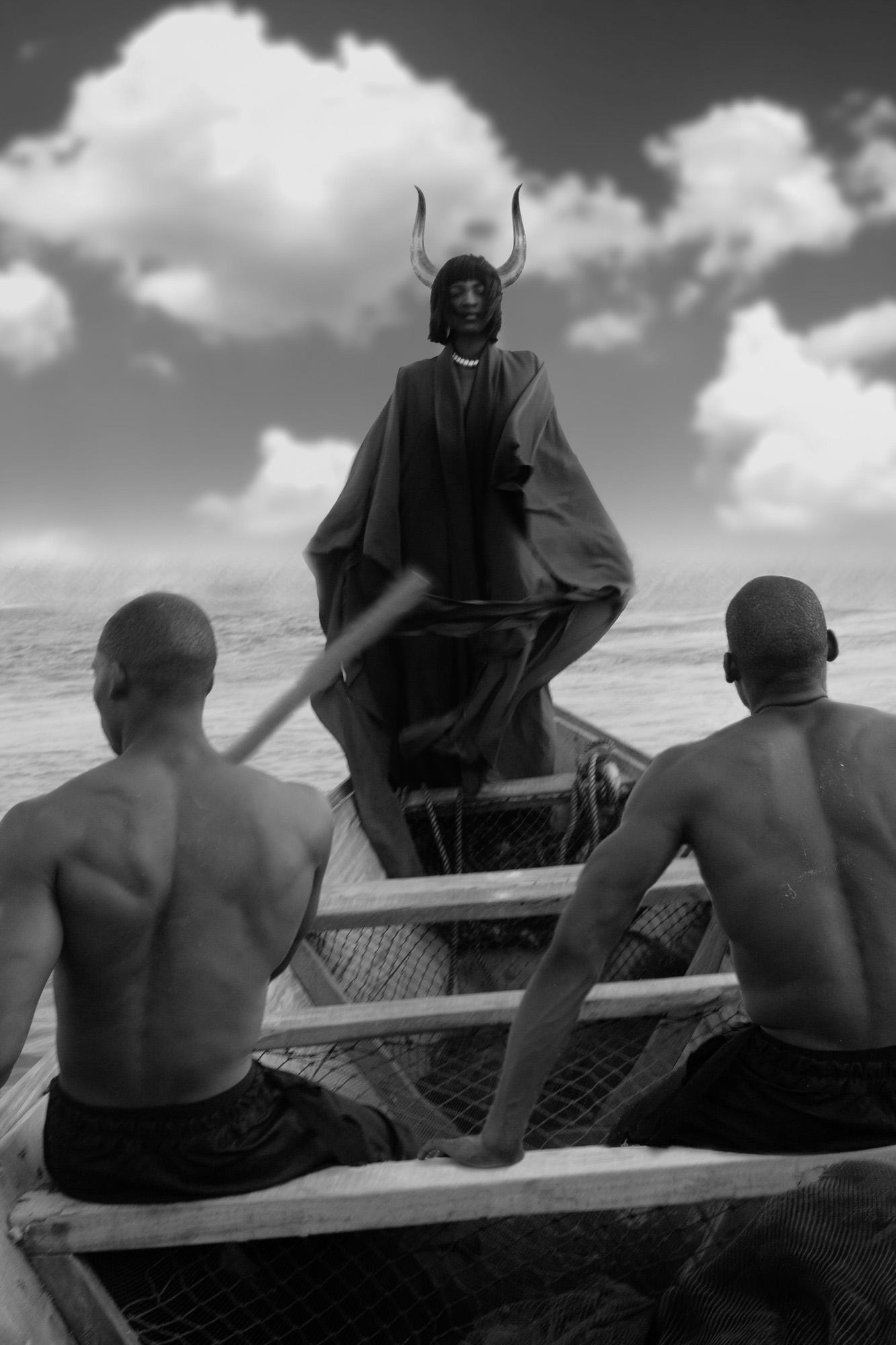 A person in a long, dark, hooded cloak with horns on their head, standing in a boat on a body of water, with a person in the front of the boat rowing.
