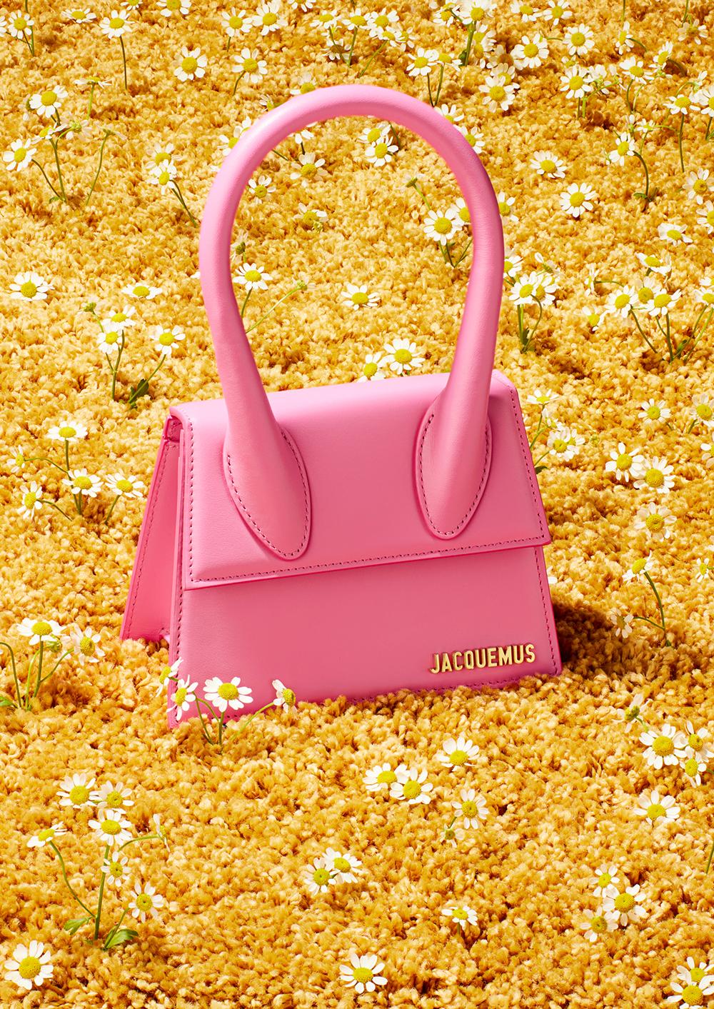 A pink handbag from the fashion brand Jacquemus, lying on a bed of yellow flowers and small white daisies.