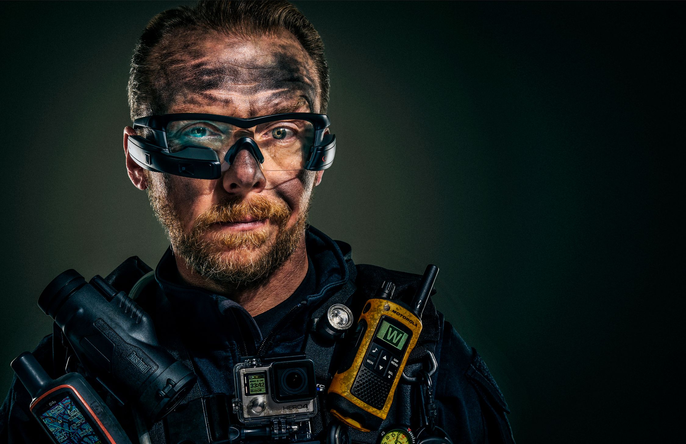 A man wearing a black jacket, bearded, with tattoos, holding a camera and various gadgets.