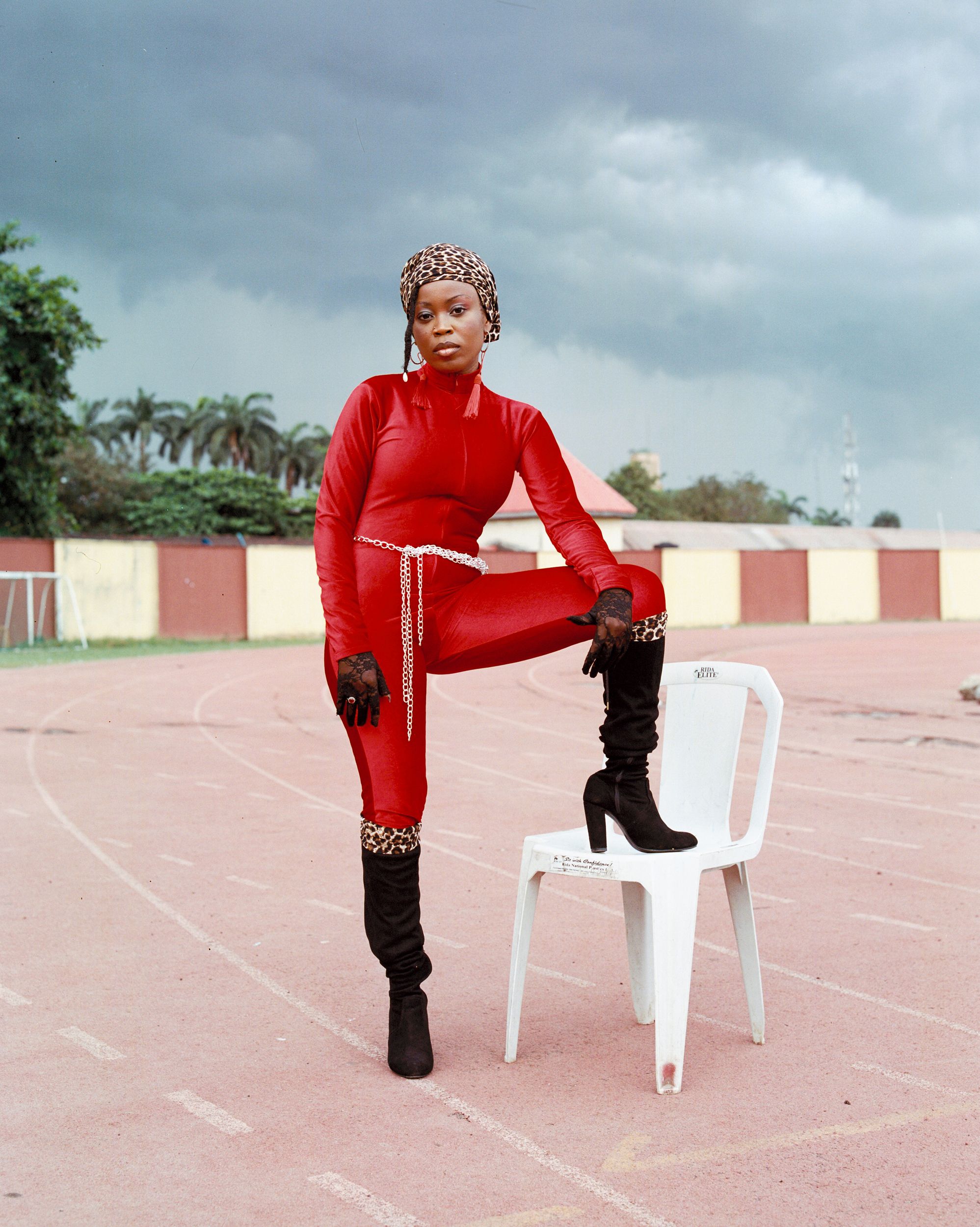 A woman poses on a track with a white chair wearing a red jumpsuit and leopard-print headscarf.