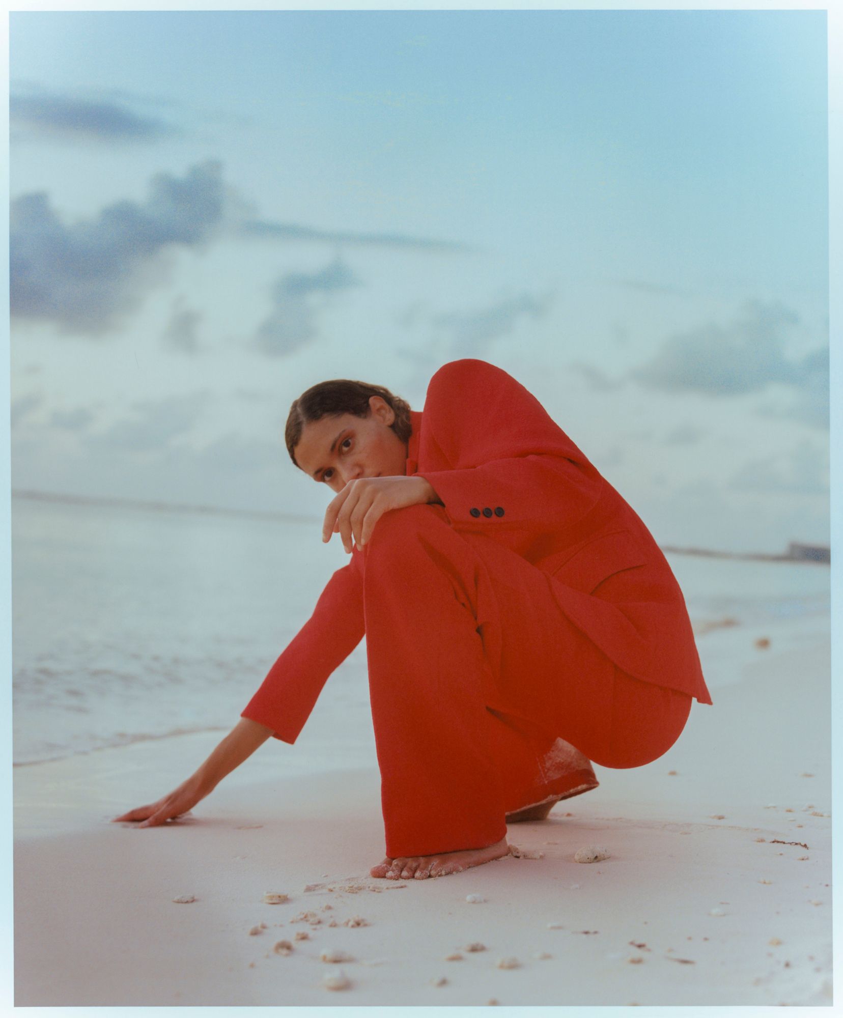 A woman in a red business suit is crouched on a beach, with her hand in the sand and her head down, looking at something in the sand.
