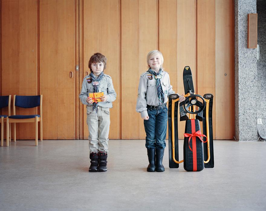 Two boys in Boy Scout uniforms hold a gift card and a snowboard in a room with a wooden wall and a chair.