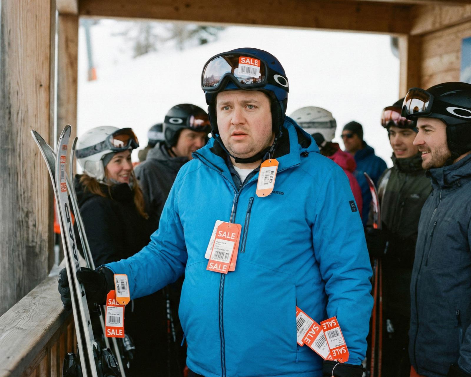 A group of people, mostly wearing winter sports gear, including helmets, jackets, and skis, standing in a snowy outdoor setting.