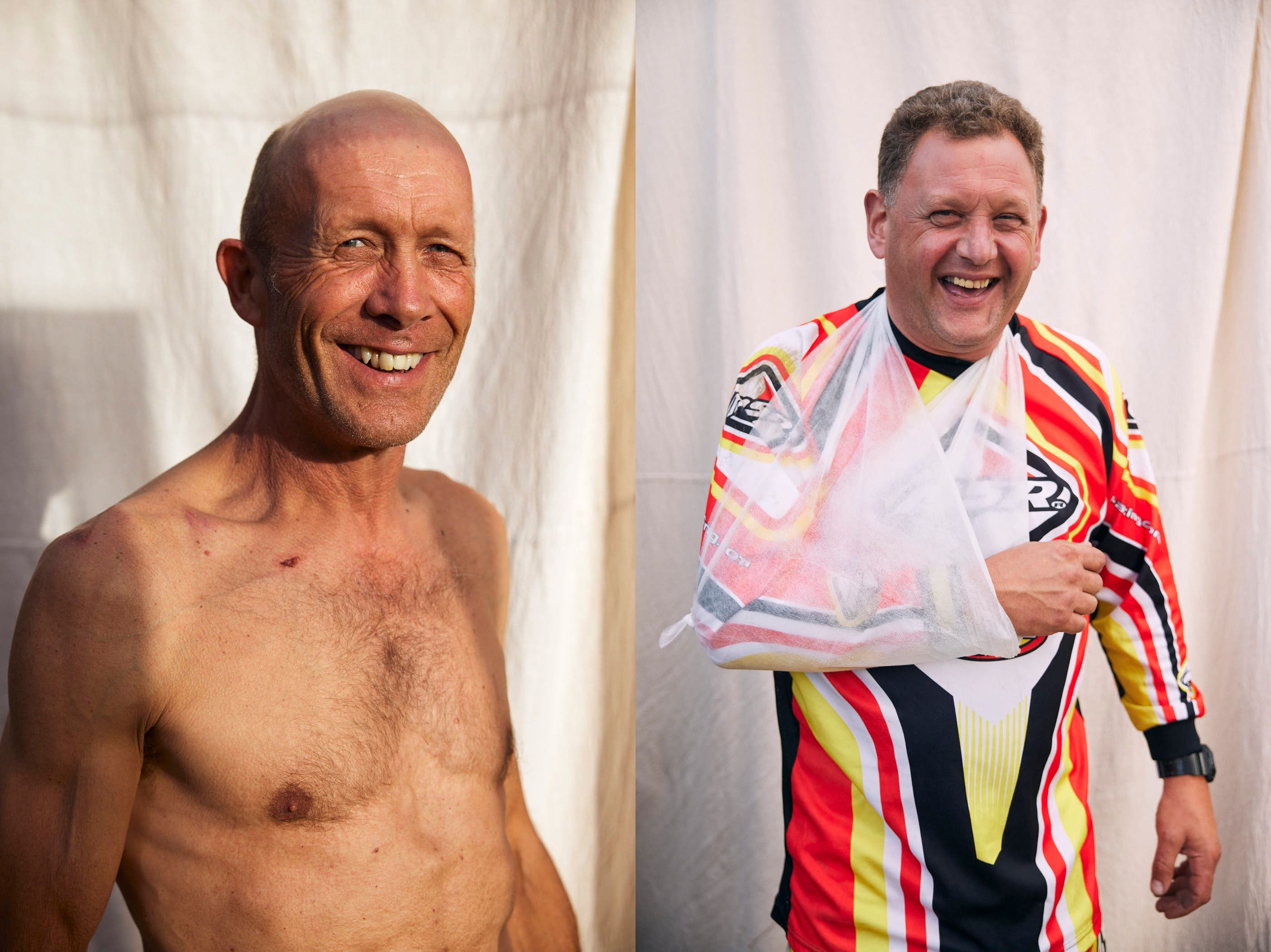 Two older men, one with a bald head and one with short hair, both with broad smiles, standing in front of a white background. The man on the left is shirtless, while the man on.