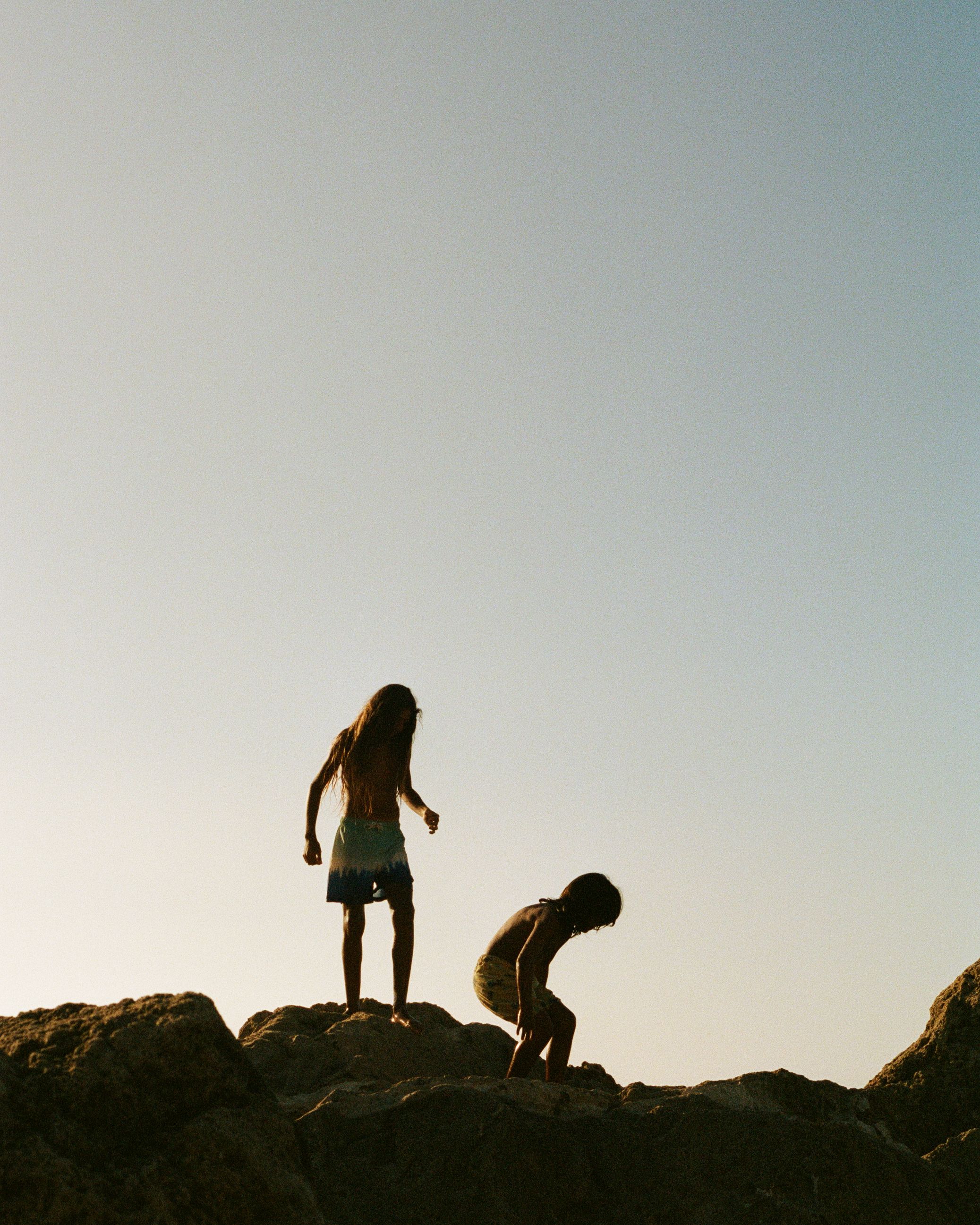 Two young children, likely siblings, are standing on a rocky outcropping, looking out at the ocean.