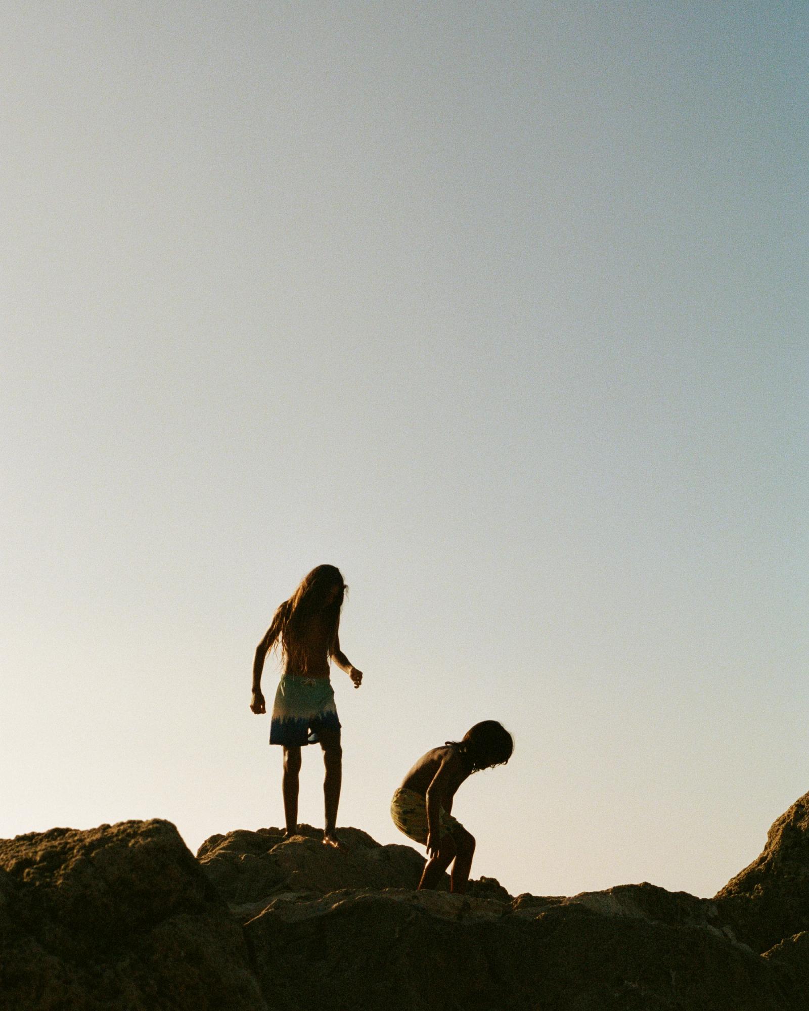 Two young children, likely siblings, are standing on a rocky outcropping, looking out at the ocean.