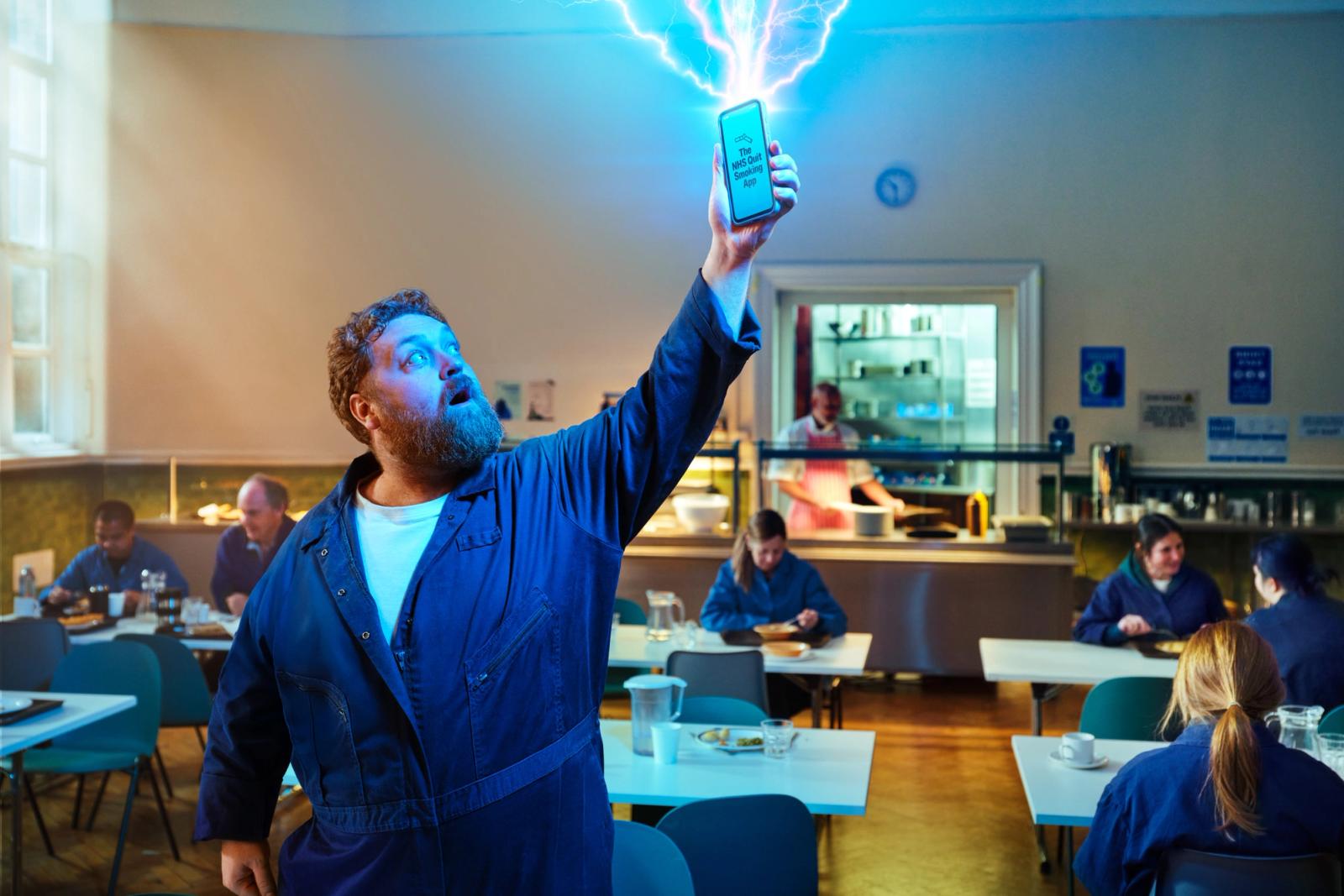 The image shows a man with a beard standing in a brightly lit cafeteria or breakroom. He is wearing blue work coveralls over a white t-shirt. He is looking up with an amazed expression at a smartphone held high in his hand, which shows "The NHS Quit Smoking App" and features glowing blue energy bolts shooting out from the screen.