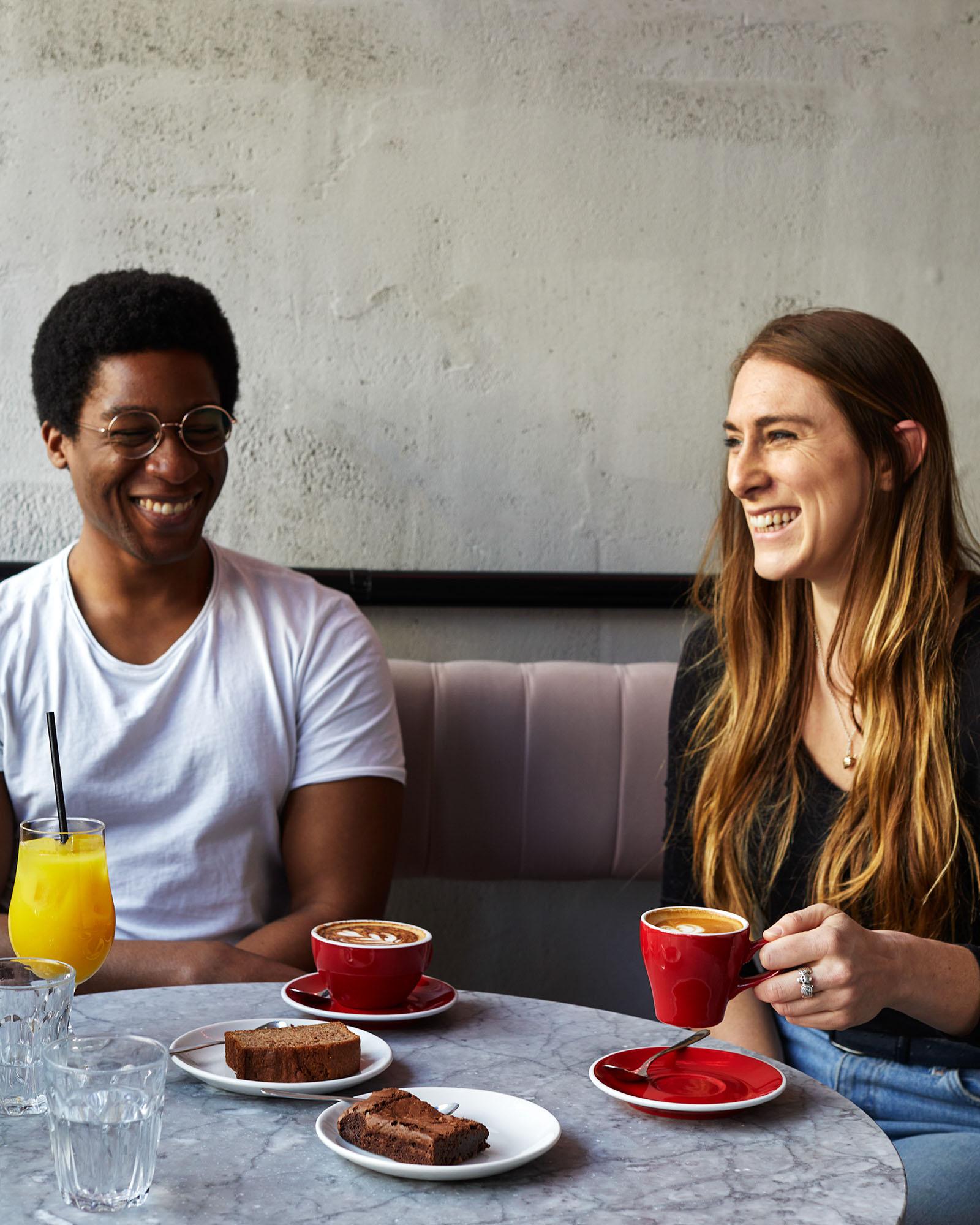 Two people sitting at a table laughing and smiling with various food and drinks including orange juice, cake, and coffee.