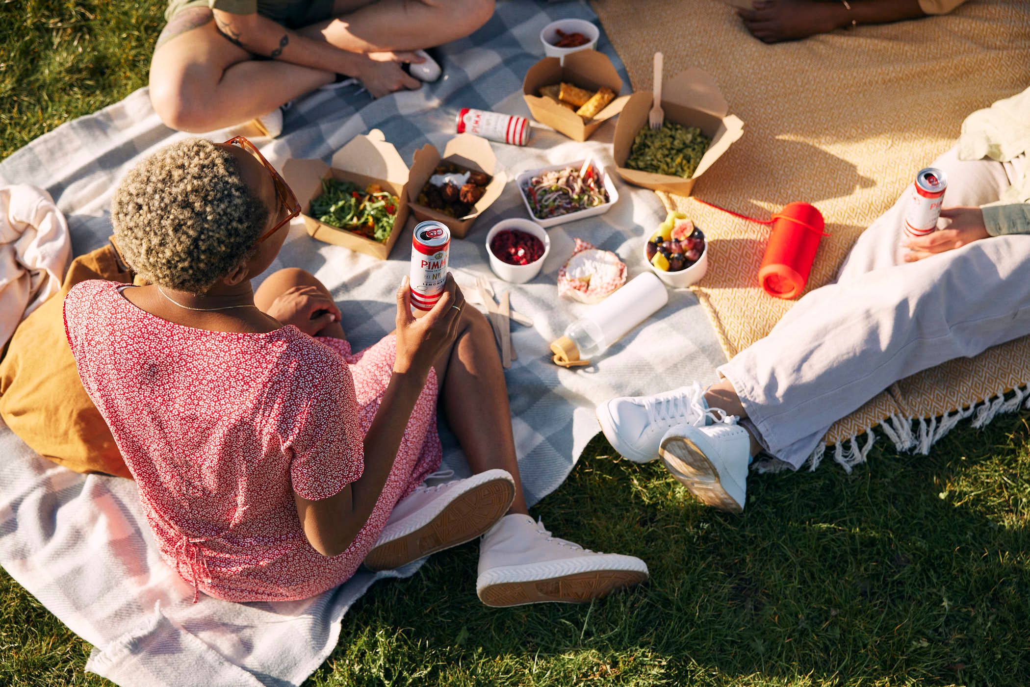 A group of people enjoying a picnic on a blanket in a park. They are surrounded by food and drinks, and everyone is smiling and laughing.