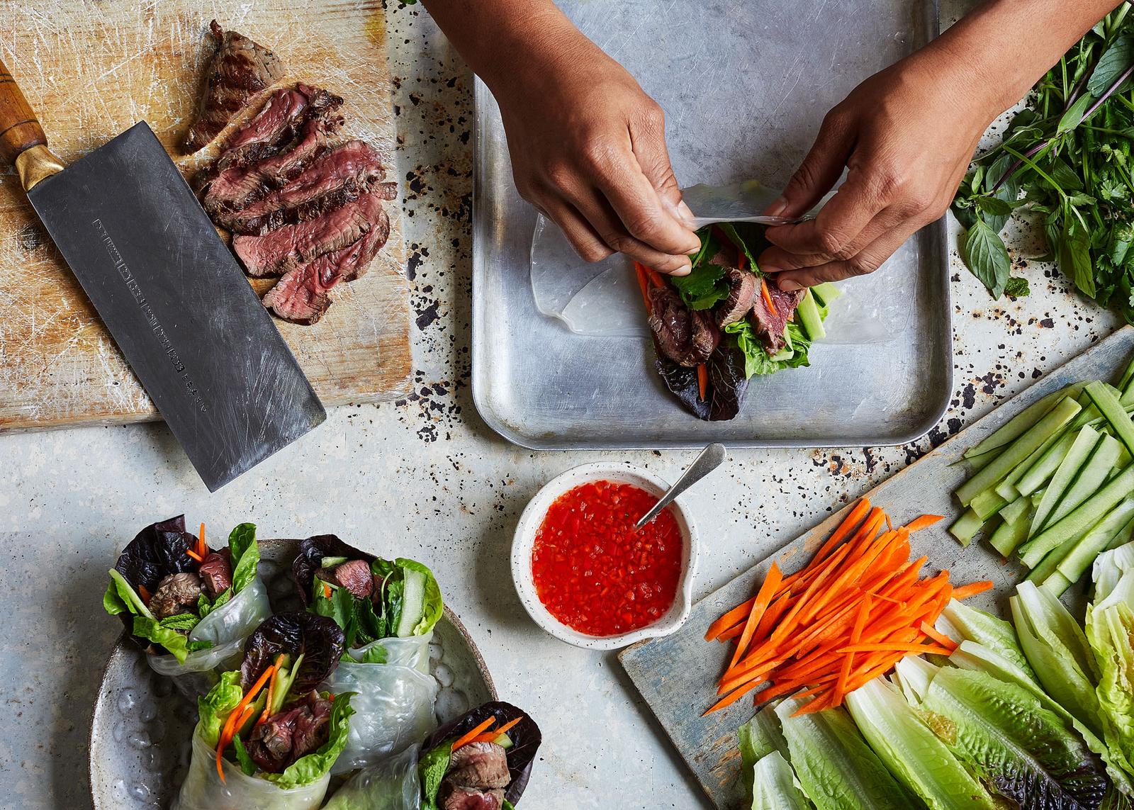 A chef's hands are preparing a dish with a steak and vegetables, with a large knife and a plate of finished food in the background.