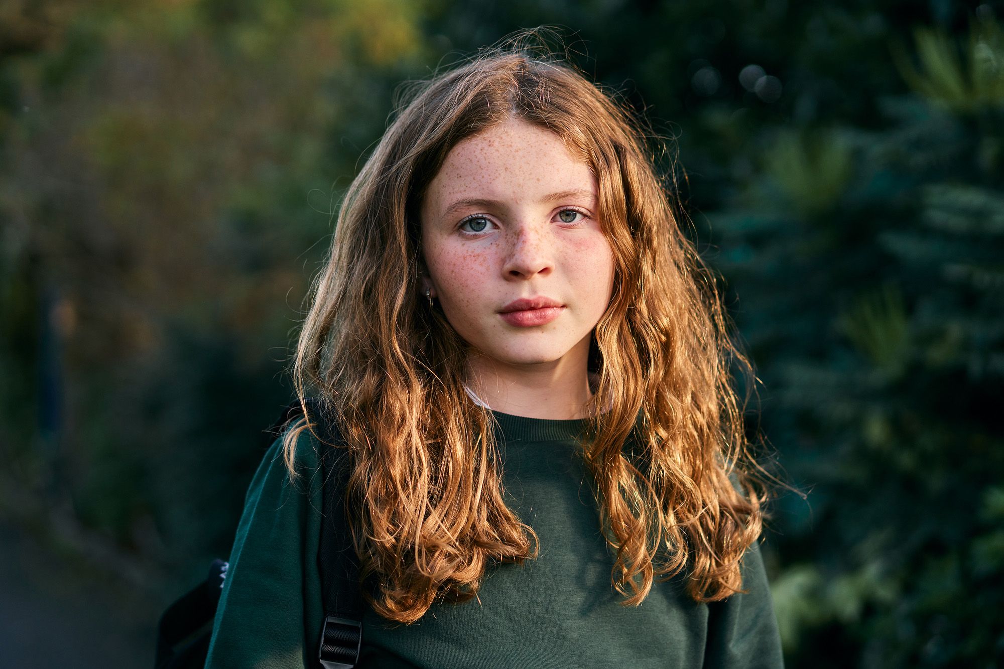 A young girl with long, curly brown hair and freckles, wearing a green sweater and standing in front of a blurred green background.