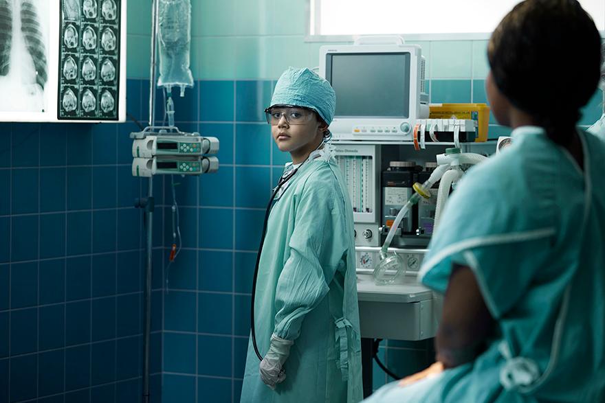 A young boy in a blue surgical gown, cap, and mask stands near medical equipment in a hospital operating room.
