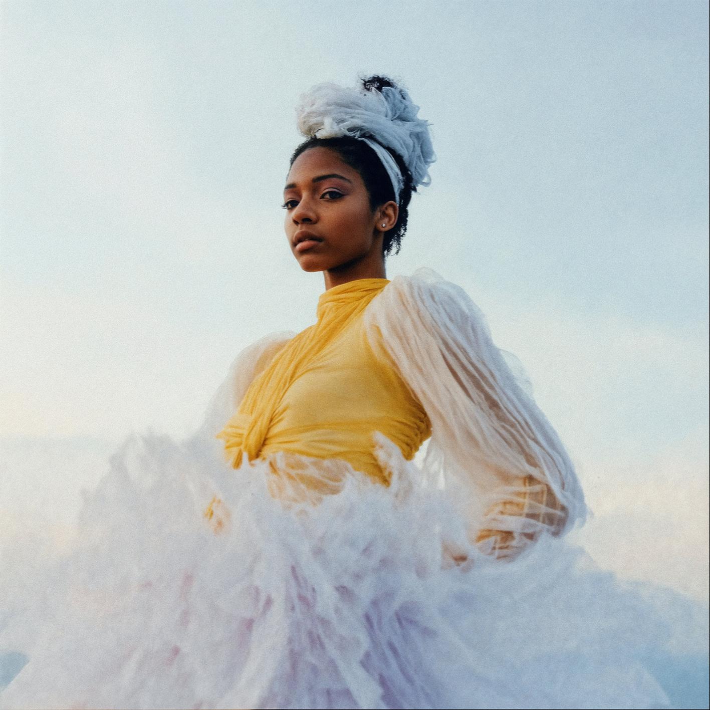 A woman in a white and yellow outfit with a large, fluffy white skirt and a matching white headband, standing in front of a cloudy sky.