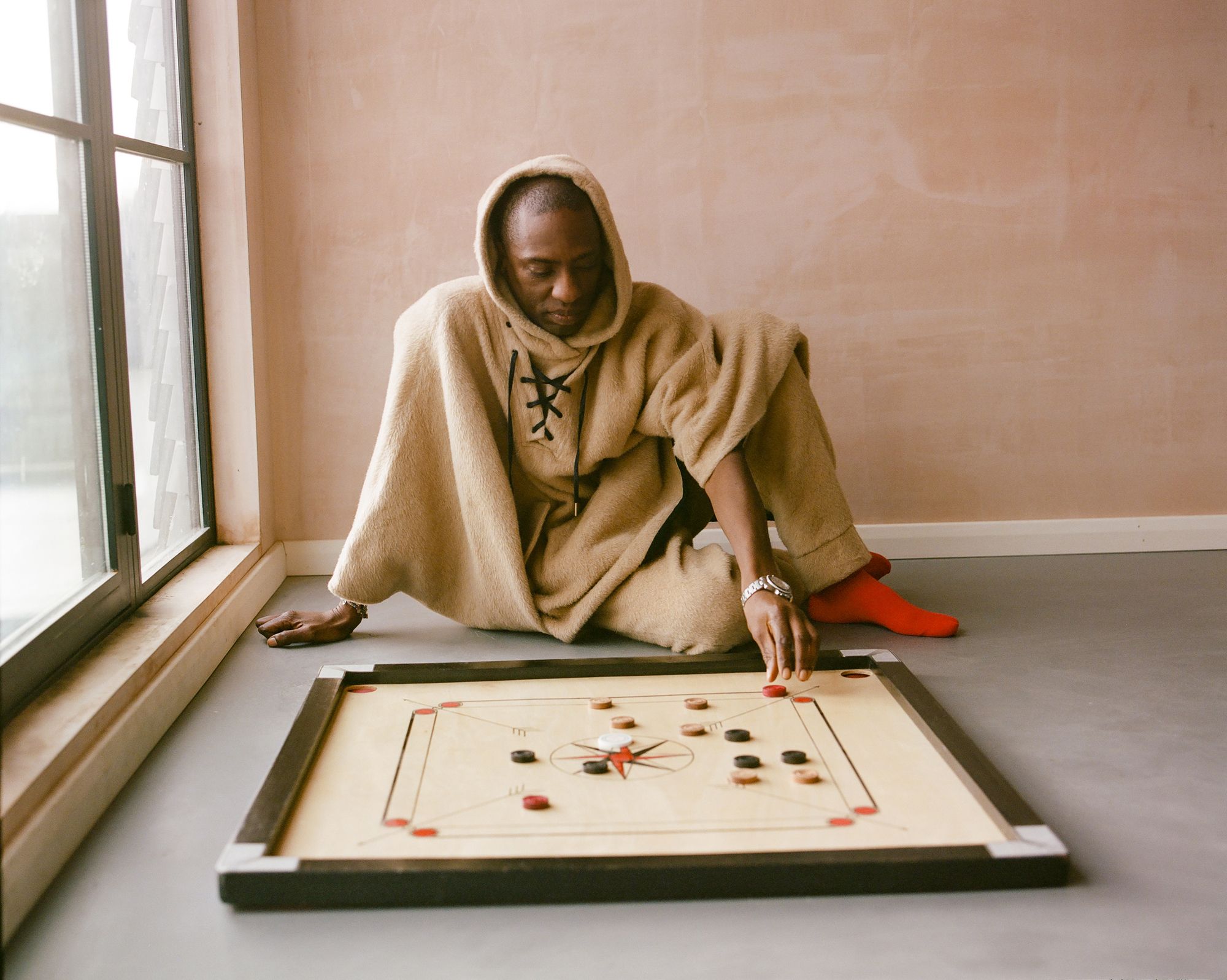 A man in a beige hooded cloak and red socks sits on the floor, playing a game of Carom (also known as Carom or Billiards) on a large, flat, wooden board.