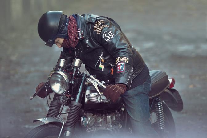 A man wearing a black leather jacket with various patches and a helmet, riding a motorcycle on a road.