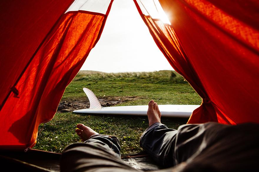 A person is lying in a tent, with a surfboard visible through the open door, in a grassy outdoor setting.