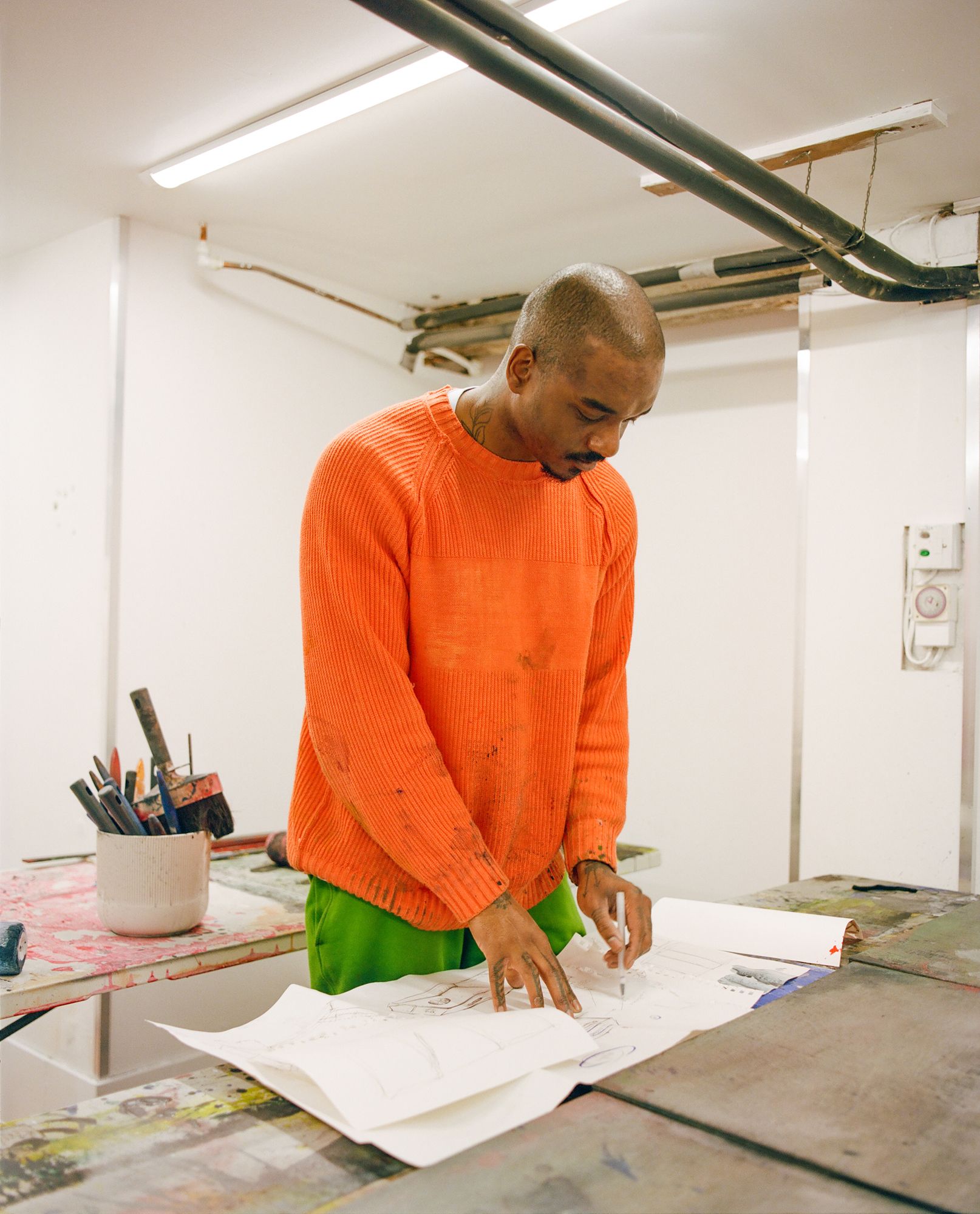 Artist Kemka Ajoku in a studio, wearing an orange sweater and green pants, standing over a table with papers and art supplies.