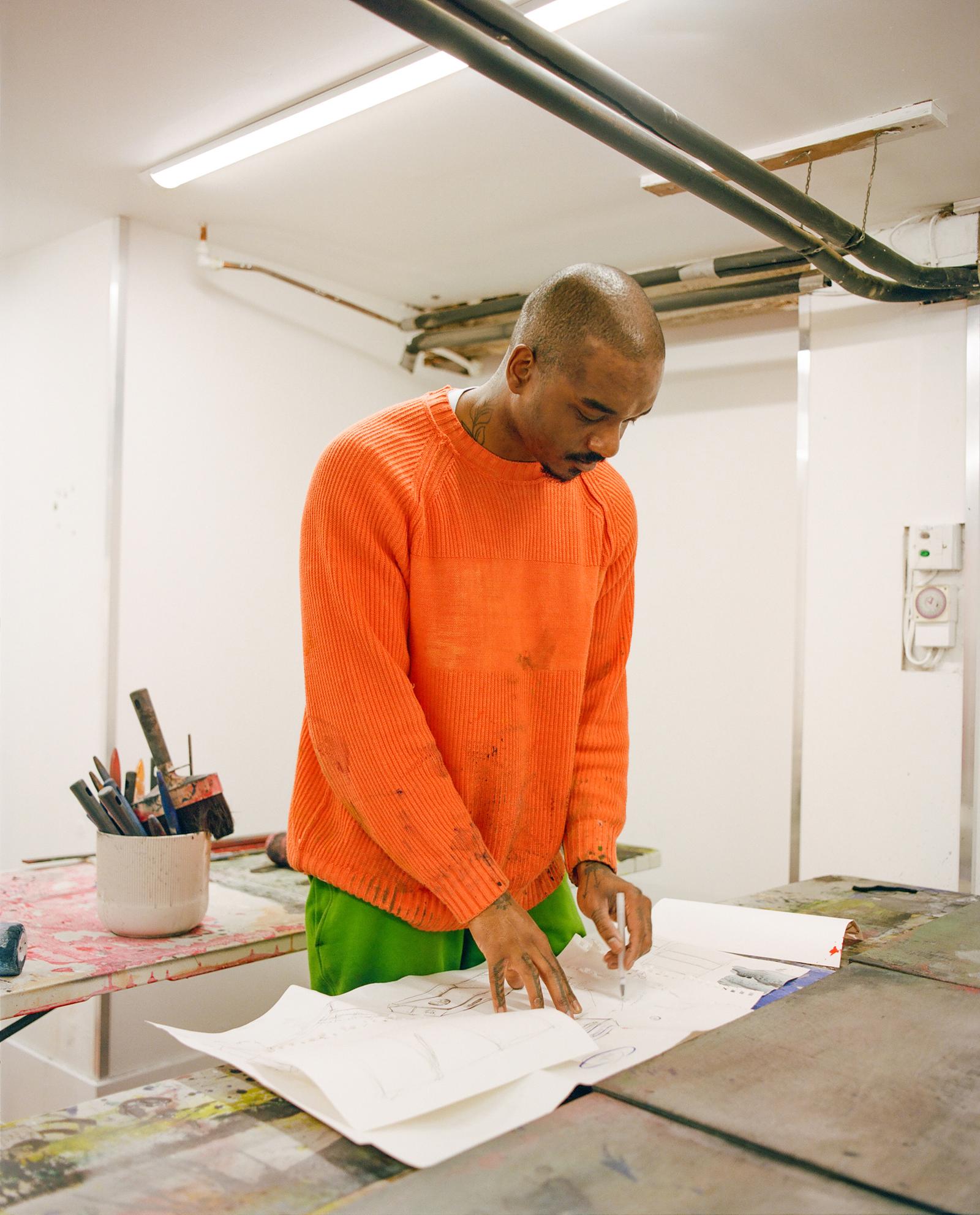 Artist Kemka Ajoku in a studio, wearing an orange sweater and green pants, standing over a table with papers and art supplies.