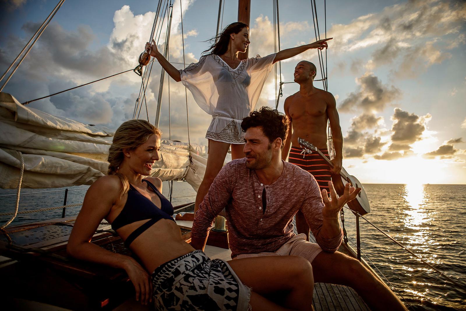 A group of people, including a woman in a white dress and a man in a red shirt, are sailing on a boat in the ocean, with a man in the background holding a surfboard.