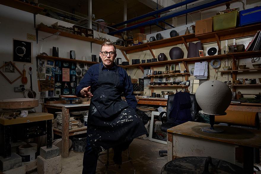 A man in a dark blue apron and shirt, sitting in a pottery studio, surrounded by various pottery tools and materials.