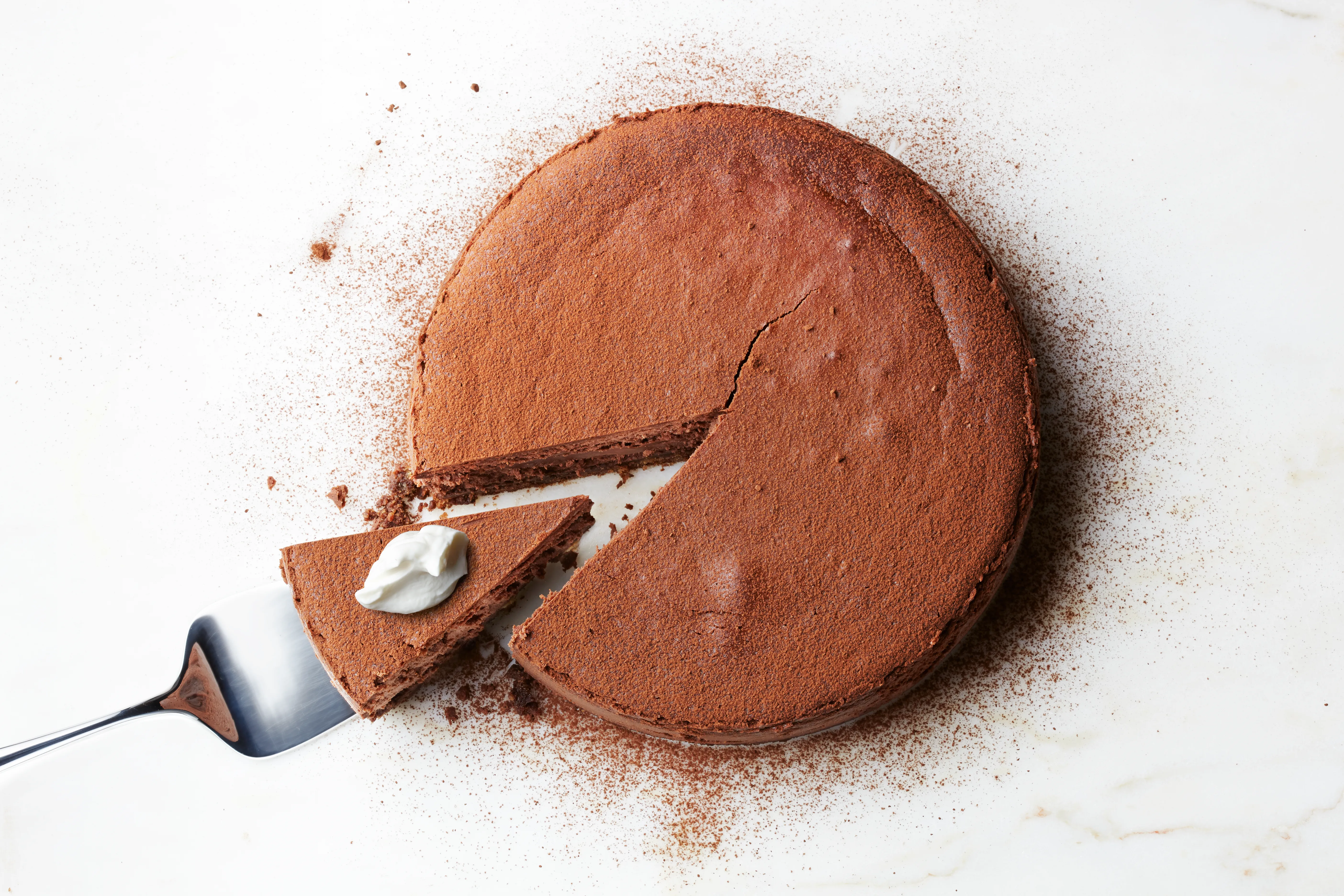 A chocolate cake with a slice removed, topped with a dollop of whipped cream, on a white marble surface.