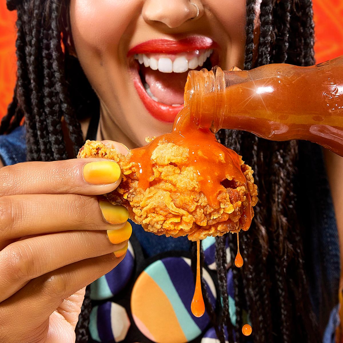 A woman with long, dark hair and a bright smile is holding a fried chicken wing and pouring a bottle of sauce over it.