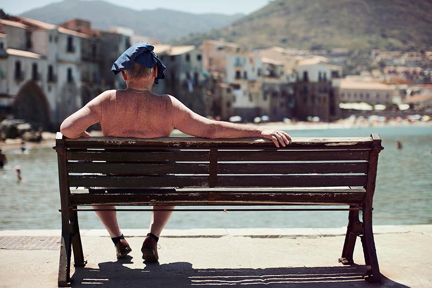 A man in a blue hat and no shirt is sitting on a bench overlooking a body of water, with a town in the background.