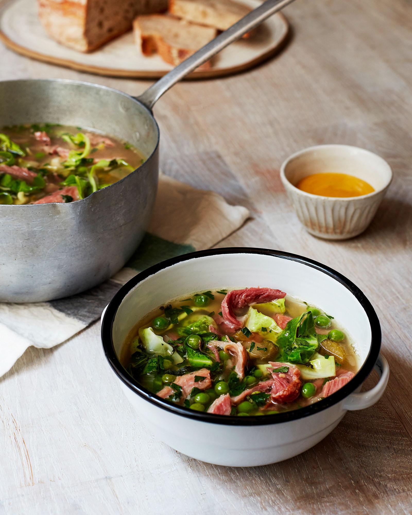 A hearty soup in a white ceramic bowl with a slice of bread and a small bowl of butter on a wooden table.